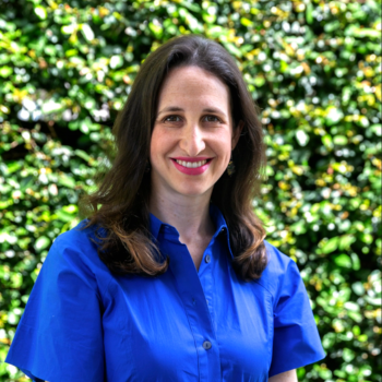 Woman in blue shirt smiling outdoors against leafy green background